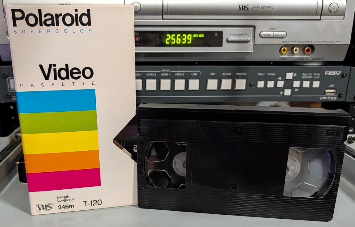 Black VHS cassette beside a white Polaroid Supercolor T-120 sleeve.