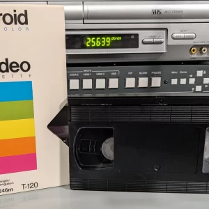 Polaroid VHS sleeve and partially obscured tape on a shelf; blue handwritten note cropped out.